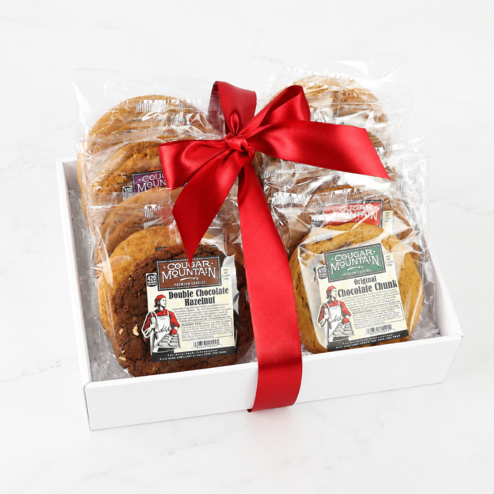 A white gift box containing assorted cookies from Cookie Mountain, wrapped in clear cellophane and tied with a red satin ribbon, on a white marble background.