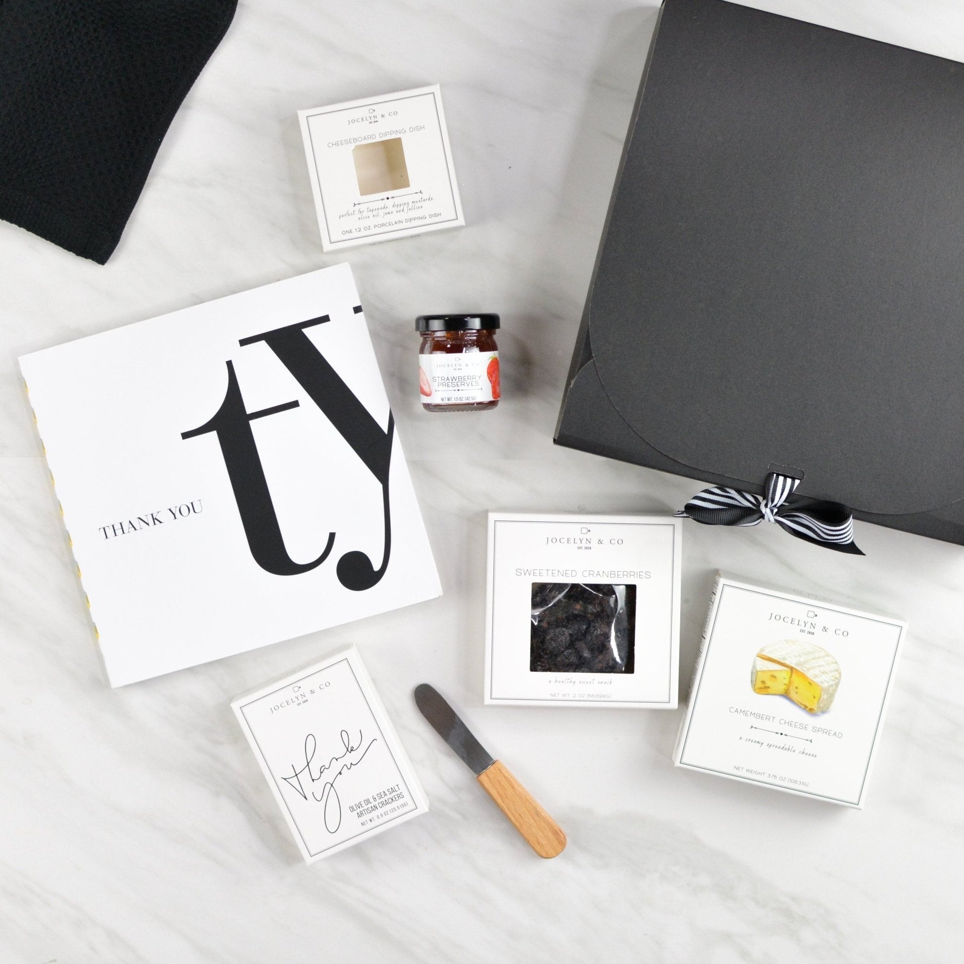 Black thank-you gift box with gratitude book, gourmet crackers, cheese, jam, and wooden spreader arranged neatly on a white surface