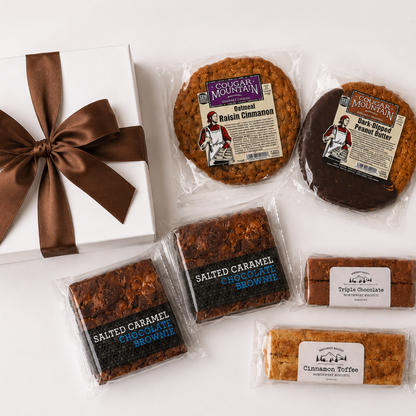 A gift assortment of cookies and brownies from Cookie Monster's Mountain, including packaged salted caramel chocolate brownie, salted caramel chocolate cookie, salted cinnamon cinnamon, and triple chocolate cookie, arranged on a white surface next to a white gift box with a brown ribbon.