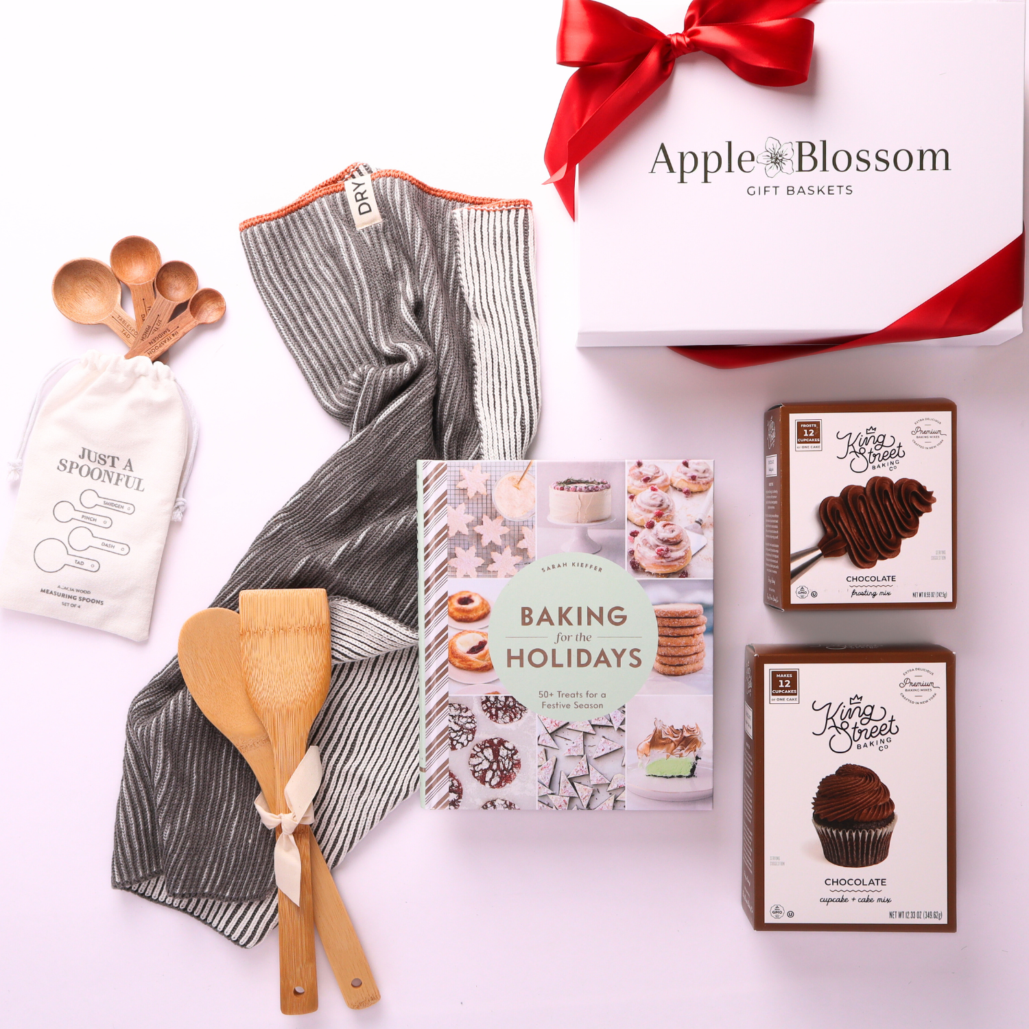 Flat lay of holiday baking gift box with Baking for the Holidays cookbook, chocolate cake mix, frosting mix, kitchen towel, wooden utensils, and measuring spoons, with a 5-star customer review.