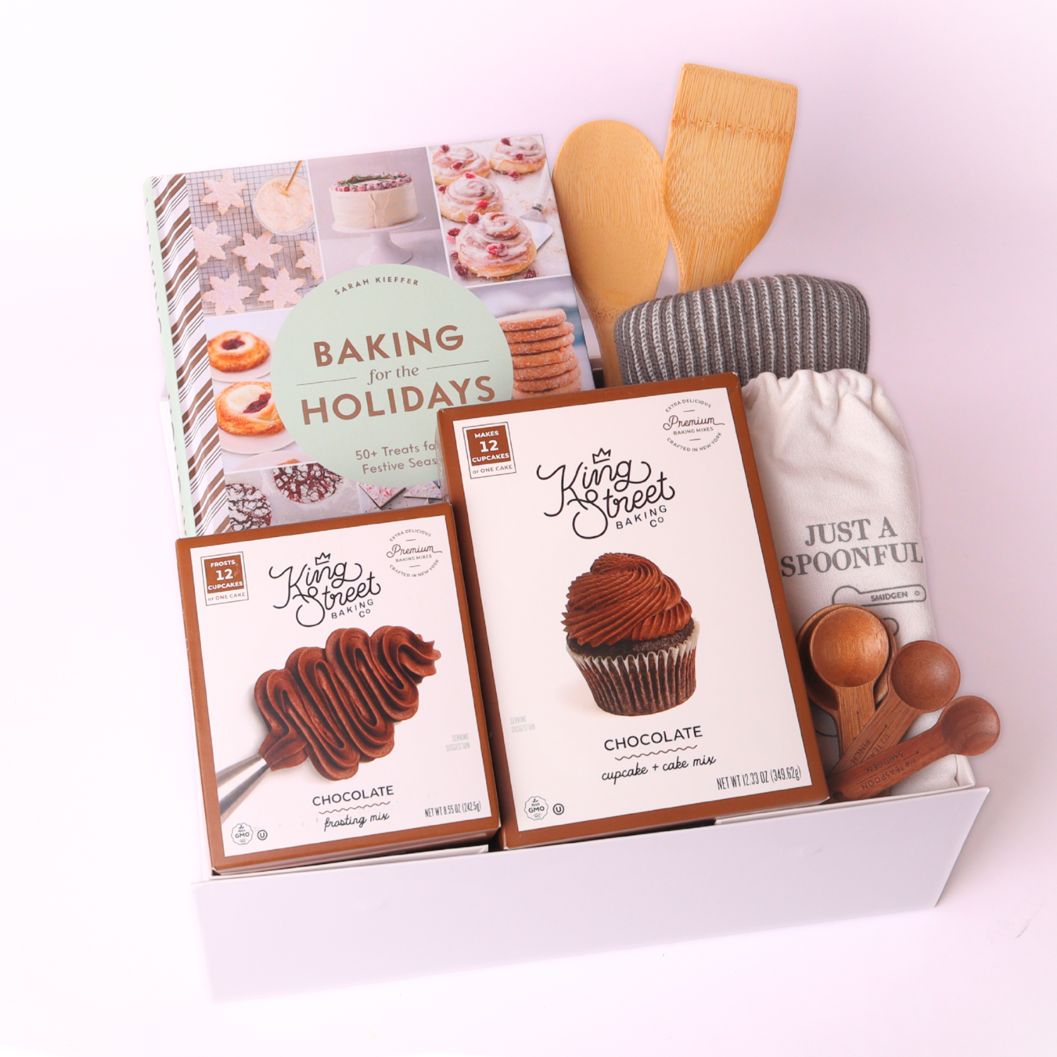 Overhead layout of Baking for the Holidays gift set featuring a cookbook, chocolate mixes, bamboo spatulas, striped dish towel, and “Just a Spoonful” spoon set next to a white gift box with red satin bow.