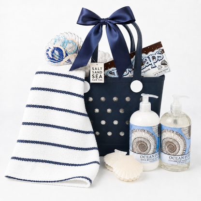 A nautical-themed gift basket set featuring a navy blue tote bag with a large navy bow, a white hand towel with navy stripes, two bottles of Ocean Reef hand wash, a seashell, and a Sea Salt & Sea candy bar, all arranged on a clean white background.