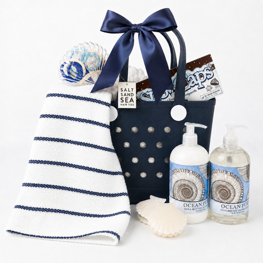 A nautical-themed gift basket set featuring a navy blue tote bag with a large navy bow, a white hand towel with navy stripes, two bottles of Ocean Reef hand wash, a seashell, and a Sea Salt & Sea candy bar, all arranged on a clean white background.