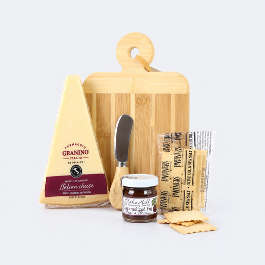A gourmet cheese and crackers gift set arranged on a light gray background, featuring a wedge of Parmigiano d'Orino Italian cheese, a bamboo cutting board, a small cheese knife, a jar of prosciutto crudo, packaged prosciutto, and flatbread crackers.