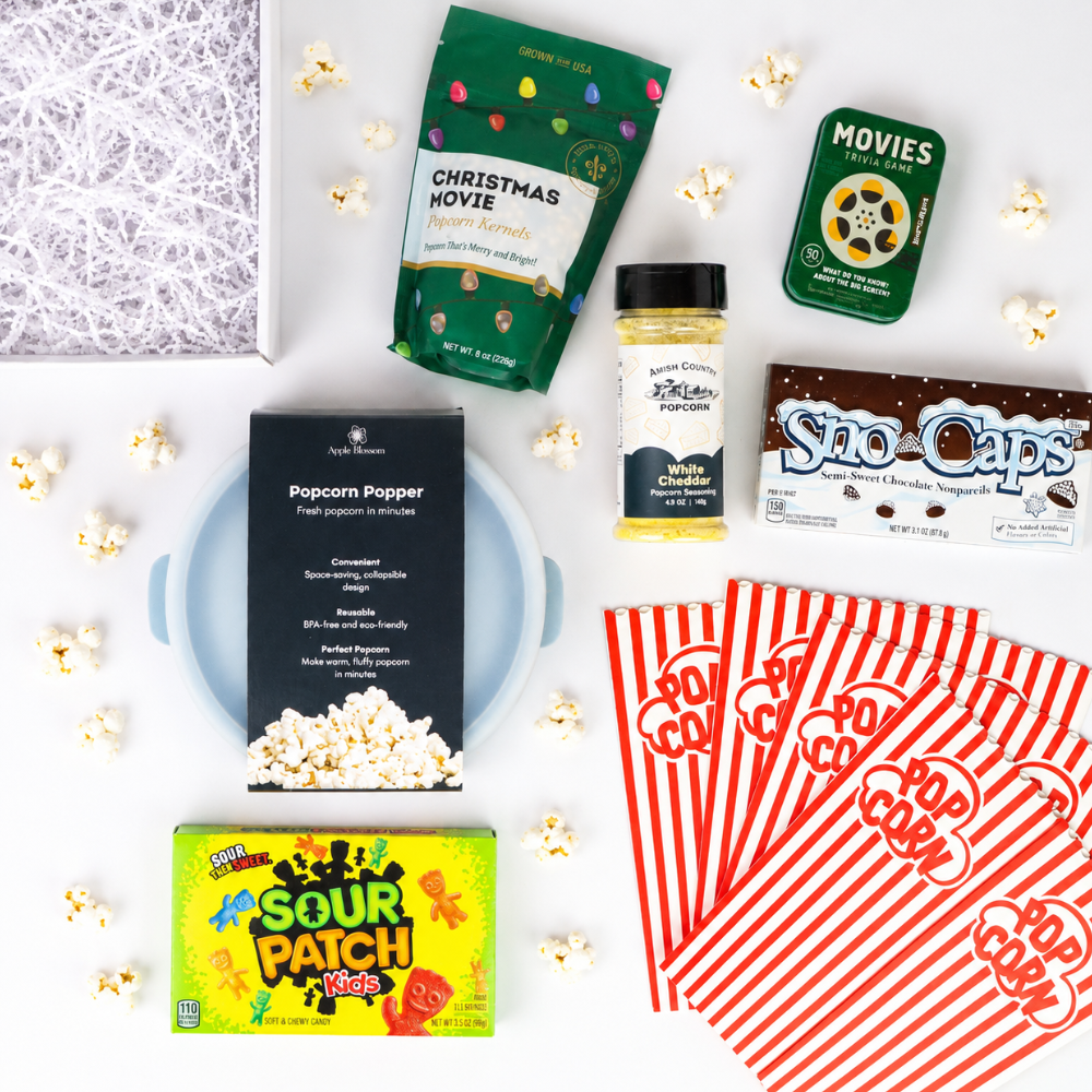 A curated movie night snack box flat lay on a white background, featuring a Popcorn Popper box, Sour Patch Kids candy, a bag of Christmas Movie popcorn, a bottle of cheese dip, Sno-Caps, red-and-white striped popcorn bags, and scattered popcorn kernels.