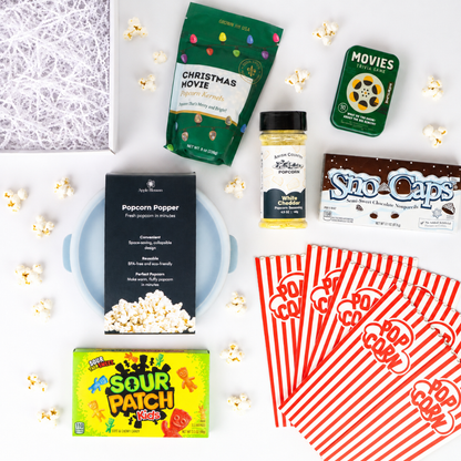 A curated movie night snack box flat lay on a white background, featuring a Popcorn Popper box, Sour Patch Kids candy, a bag of Christmas Movie popcorn, a bottle of cheese dip, Sno-Caps, red-and-white striped popcorn bags, and scattered popcorn kernels.