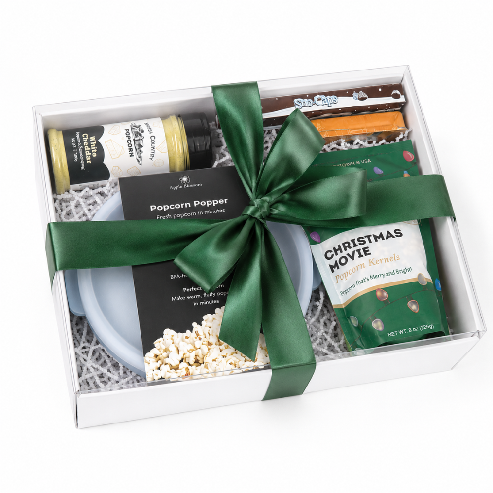A curated gift box set containing a Popcorn Popper bottle, a green bag of Christmas Movie popcorn, a blue bowl, and assorted chocolates, wrapped in a dark green satin ribbon.