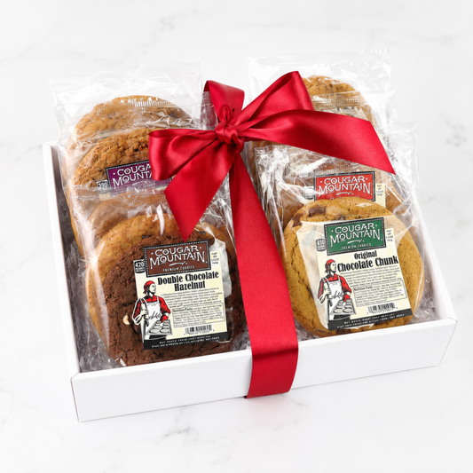 A gift box of Cougar Mountain cookies on a white marble surface, featuring two packages of cookies tied together with a red satin ribbon. The packages show labels for Cougar Mountain Double Chocolate Hazelnut and Cougar Mountain Original Chocolate Chunk cookies.