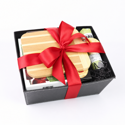 A black gift box containing a bamboo cutting board, a red satin ribbon, and a small packaged item, photographed on a white background.