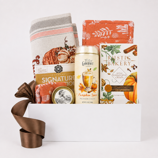 A curated gift basket set in a white box, featuring a striped kitchen towel with a pumpkin design, a rust-orange fabric piece, a can of Toffee Gourmet Pumpkin Spice Cacao, a box of Rustis Bakery Spiced Chews, and a small spice tin, styled with a brown ribbon bow on a white background.
