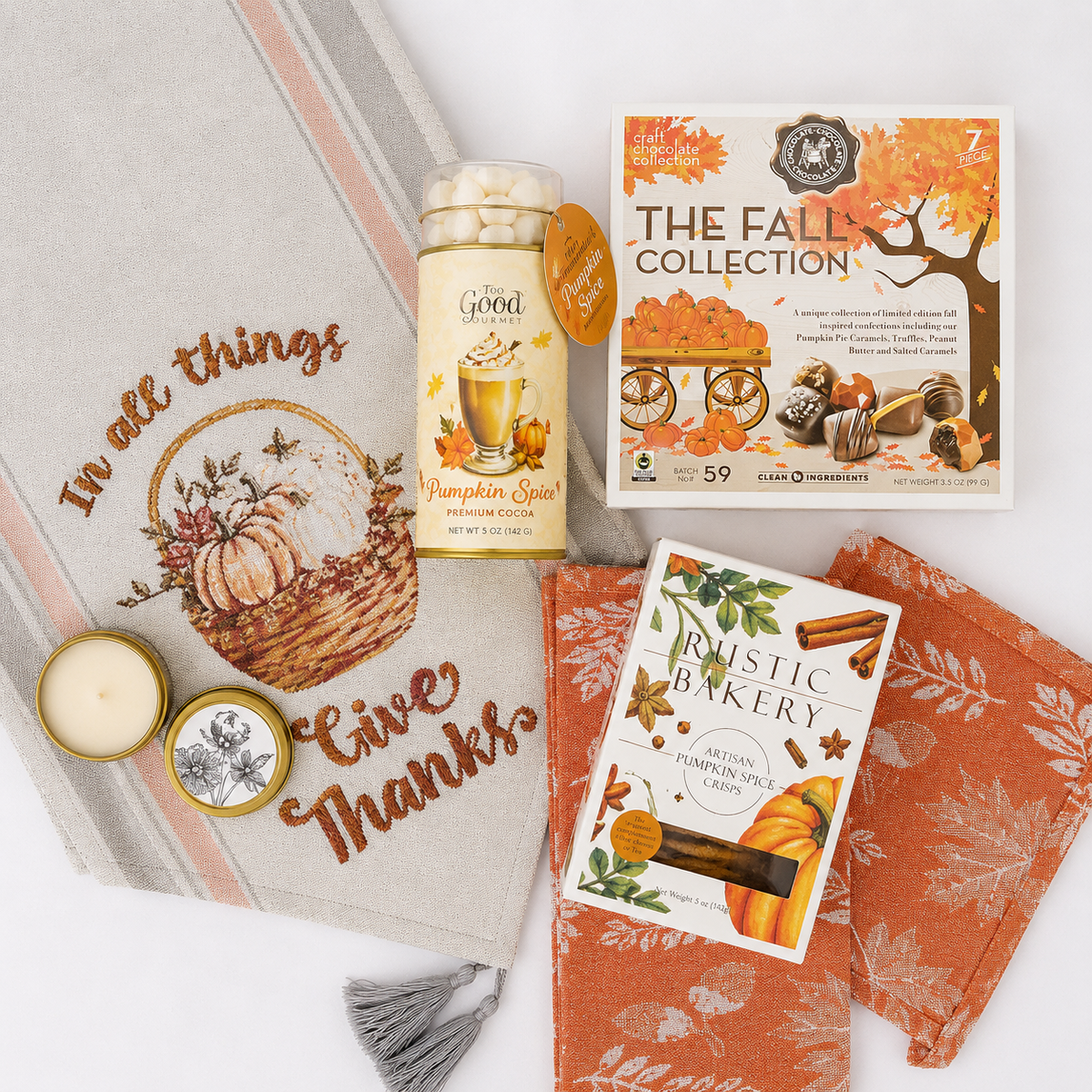 A coordinated fall-themed gift set arranged on a white background, including a beige decorative throw blanket with embroidered autumn text and pumpkin illustration, an orange patterned cloth napkin, a jar of pumpkin spice marshmallows, a box labeled The Fall Collection, and a card/booklet titled Rustic Baker Autumn Pumpkin Spice.