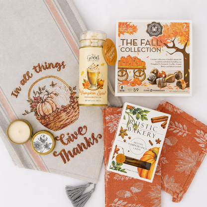 A coordinated fall-themed gift set arranged on a white background, including a beige decorative throw blanket with embroidered autumn text and pumpkin illustration, an orange patterned cloth napkin, a jar of pumpkin spice marshmallows, a box labeled The Fall Collection, and a card/booklet titled Rustic Baker Autumn Pumpkin Spice.