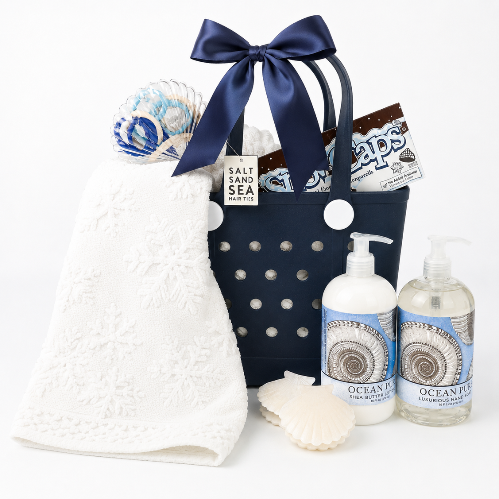 A curated bath and beach-themed gift set on a white background, featuring a navy blue tote bag with a large navy bow, a white textured towel, two bottles of Ocean Life hand and body products, a wrapped candy, and seashell props.