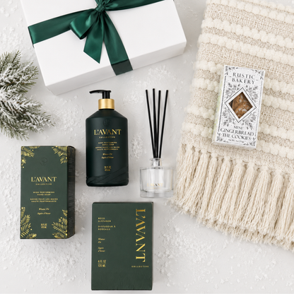 A curated gift set featuring L'AVANT products, including a dark green gift box with a green ribbon, a black body mist bottle with a gold cap, a reed diffuser with black reeds, and a dark green box, arranged on a white textured surface next to a cream knit blanket and a small evergreen sprig.