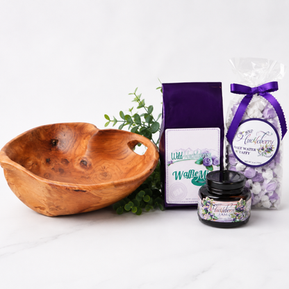 A gift set featuring a carved wooden bowl, a purple bag of waffle mix, a jar of huckleberry waffle mix, and a bag of lavender salt water tea puffs on a white marble surface.