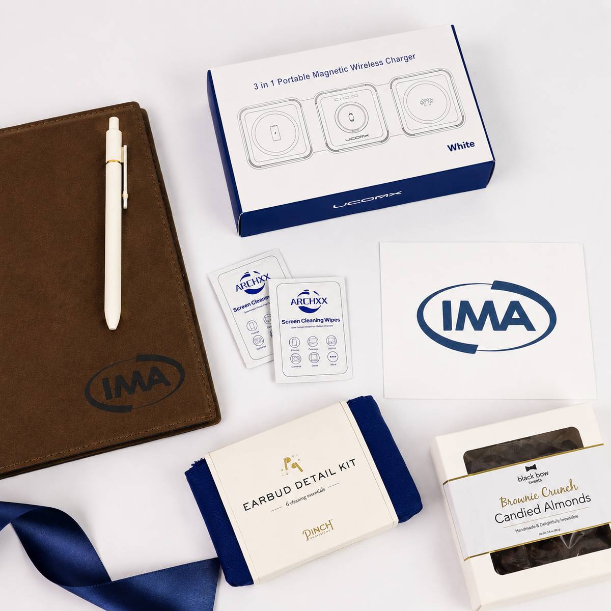 Collection of IMA branded items including a notebook, pen, magnetic wireless charger, earbud detail kit, and candy on a white background.
