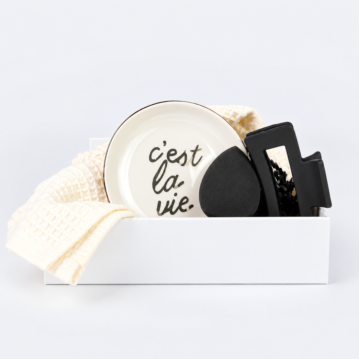 A white ceramic plate with the French phrase 'c'est la vie.' in black script, styled in a gift box with a cream waffle-knit towel and black accessories on a white background.