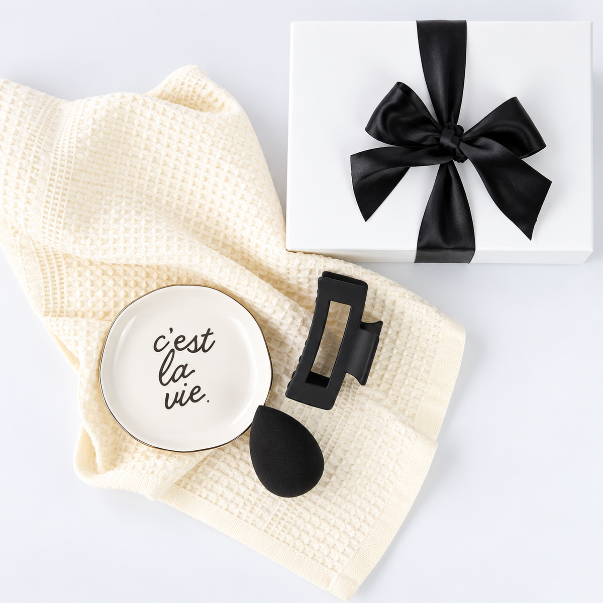 A styled flat-lay featuring a cream waffle-knit towel, a small white ceramic dish with gold trim and the French phrase 'c'est la vie.' in black script, a black hair claw clip, and a white gift box tied with a black satin bow on a white background.