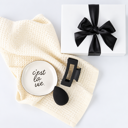 A styled flat-lay featuring a cream waffle-knit towel, a small white ceramic dish with gold trim and the French phrase 'c'est la vie.' in black script, a black hair claw clip, and a white gift box tied with a black satin bow on a white background.