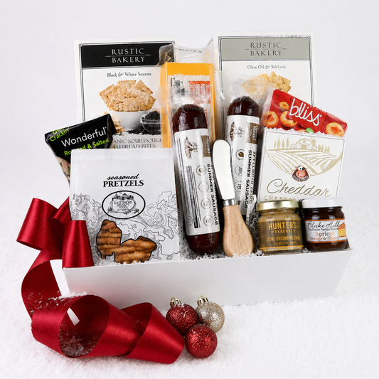 A curated gourmet gift basket arranged in a white box with a large red ribbon bow, featuring Rustic Bakery items, Hunter's Creamery sausage, Bliss cheddar, pretzels, and jars of honey, set on a white background with red and silver ornaments in front.