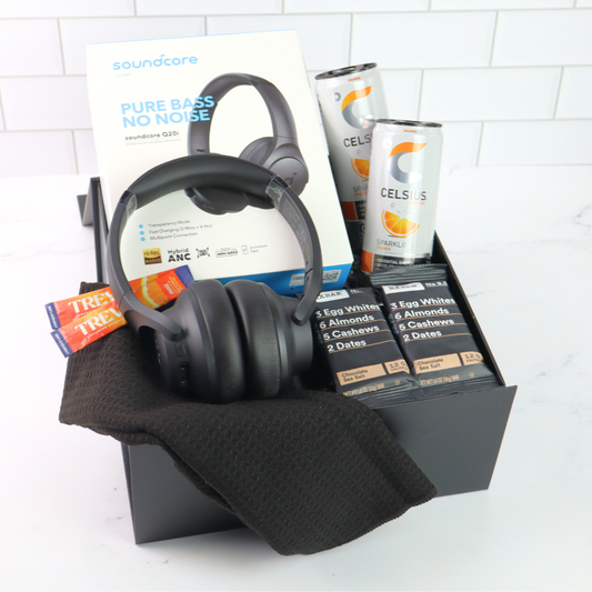 Black headphones with packaging, Celsius drinks, and protein bars in a box on a white tiled background