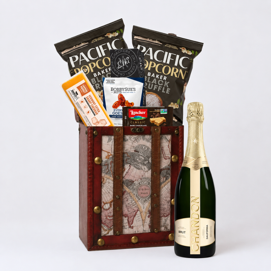 A gourmet gift basket set containing a bottle of Brut sparkling wine, bags of Pacific Popcorn and Ruffie's Black Ruffle chips, and a map-patterned wooden crate with brown leather straps.