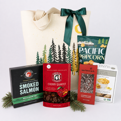 A gift basket set featuring a cream canvas tote bag with a green ribbon and pine tree print, surrounded by assorted snack boxes and bags including Trader Joe's Smoked Salmon, G. Maxwell's Cherry Quartet, and Pacific Popcorn.